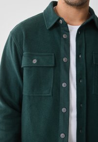 Maglia in pile verde con chiusura a bottoni, due taschini sul petto e colletto classico. I bottoni hanno contorni neri, il tessuto ha una texture morbida.