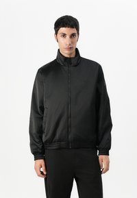 Zwarte zip-up jas van gladde stof met een hoge kraag, elastische manchetten en een slimfit tailleband. Bevat subtiele branding op de borst.