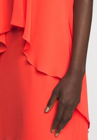 Mörkhyad hand med naturliga naglar som vilar mot en ljus korallfärgad klänning med lager och volanger.