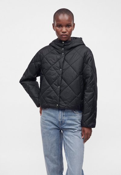 Jeune personne portant une veste matelassée noire avec capuche et un jean en denim bleu clair, posant devant un fond clair uni.