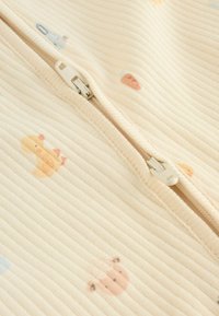 Bēšs ar rāvējslēdzēju apģērbs ar ribotu struktūru, uz auduma krāsainas dzīvnieku apdrukas, tostarp pīle, aita un burkāns.