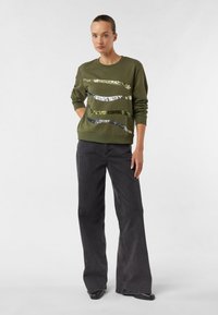 Oliwkowa bluza z cekinowymi falistymi paskami w srebrnym i złotym kolorze; zestawiona z czarnymi spodniami o szerokich nogawkach i czarnym obuwiem.