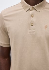 Gros plan d'une personne portant un polo beige avec trois boutons et un petit logo géométrique brodé sur la poitrine.