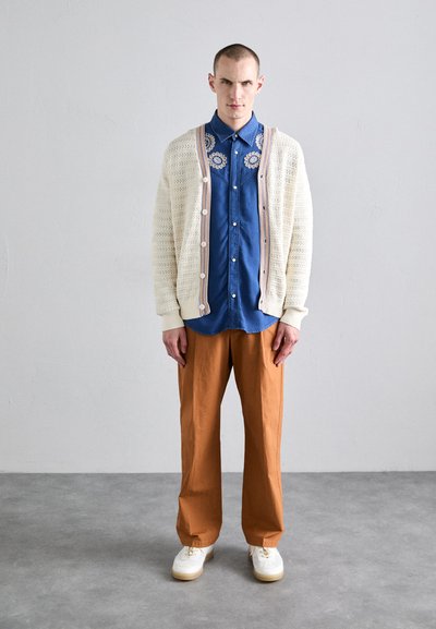 Strickjacke in Creme mit Knopfverschluss über einem blauen bestickten Hemd, kombiniert mit lockeren braunen Hosen und weißen Turnschuhen.