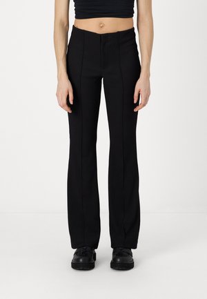 Person wearing high-waisted black fitted pants with vertical seams, black chunky shoes, and a cropped black top against a plain white background.