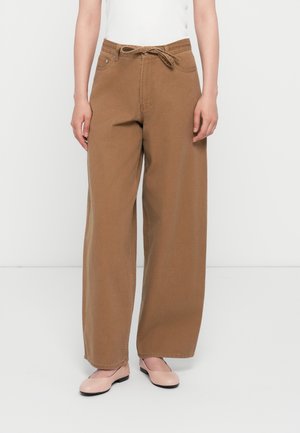 Persoon met bruine wijde broek met een gestrikte taille en lichtroze platte schoenen, staand tegen een effen witte achtergrond.