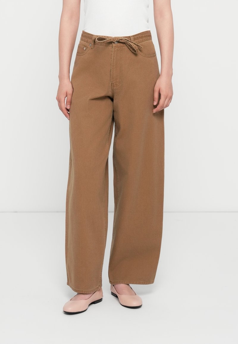 Personne portant un pantalon marron à jambes larges avec une taille nouée et des chaussures plates rose clair, debout devant un fond blanc uni.