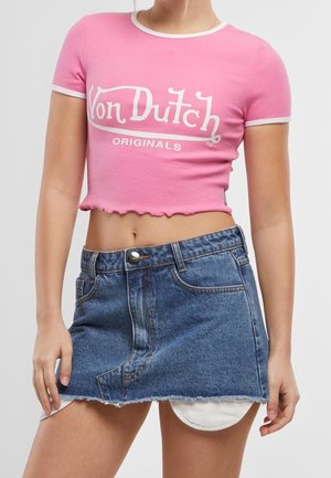 Person iført en pink Von Dutch kort t-shirt og et kort blåt denimskørt med flossede kanter over hvide shorts.