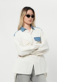Frau mit langen Haaren, die ovale Sonnenbrillen trägt, ein übergroßes cremfarbenes Hemd mit blauem Denim-Kragen und -Tasche sowie hellgraue Hosen hat und die Arme verschränkt.