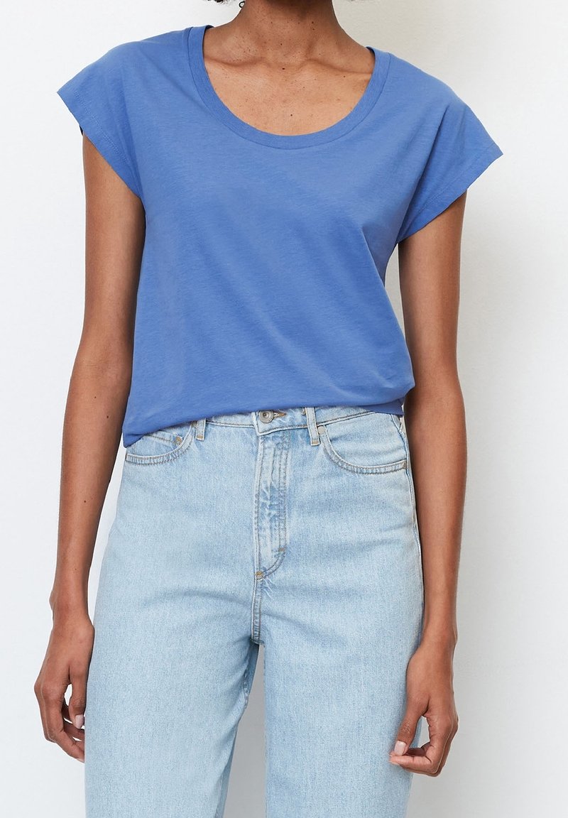 Femme portant un t-shirt bleu à manches courtes et un jean taille haute bleu clair, les mains détendues le long du corps sur fond blanc.