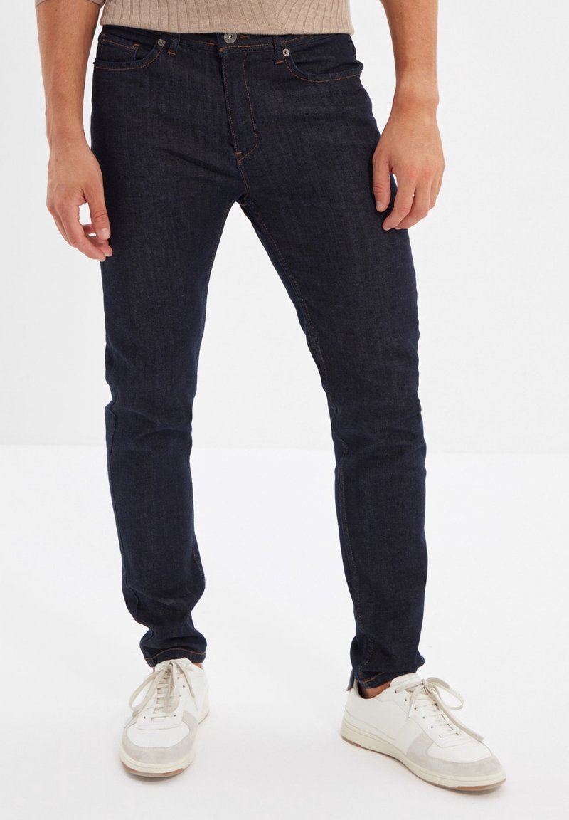 Homme portant un jean slim bleu foncé et des baskets blanches, debout devant un fond blanc uni.