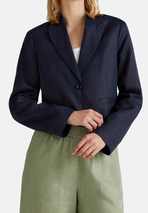 Mujer con blazer azul marino de un solo botón sobre una blusa blanca y pantalones de cintura elástica verde claro, con las manos entrelazadas a la altura de la cintura.