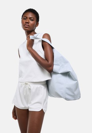 Jeune femme portant un débardeur blanc et un short, portant un grand sac cabas rayé bleu clair sur l'épaule.