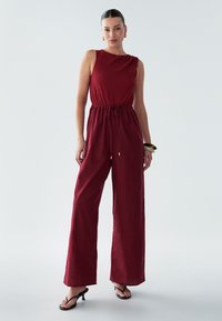 Combinaison bordeaux avec un haut sans manches texturé, taille élastique et pantalons à jambes larges, dotée d'un cordon de serrage et d'accents dorés.