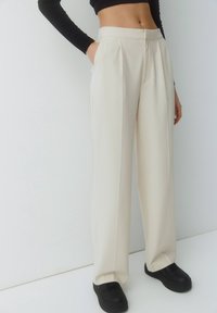 Pantalones blancos con pliegues, de cintura alta, fabricados en un tejido ligero, con bolsillos laterales y un diseño de pierna recta, combinados con zapatillas negras.