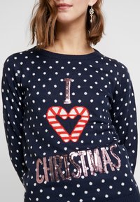 Pull marin à pois blancs, avec un design en forme de cœur en canne de Noël rouge et blanc et un texte en sequins rose doré brillant "NOËL."