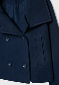 Manteau en laine bleu marine à double boutonnage avec de larges revers, quatre boutons noirs et une poche passepoilée verticale à l'avant.