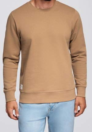 Mann trägt einen schlichten hellbraunen Rundhalspullover und hellblaue Jeans, steht mit locker an den Seiten herabhängenden Armen.