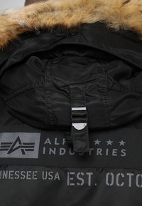 Black jacket with a faux fur-lined hood, silver hardware, and prominent branding label featuring "Alpha Industries" and establishment details.