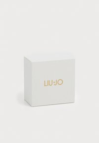 Väike valge tekstuuriga ehtekarp, mille esiküljel on kuldne LIU·JO logo, hallika tausta vastu.