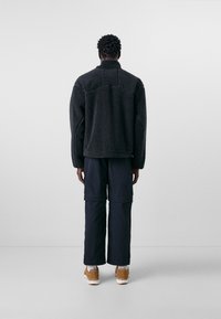 Schwarze Fleecejacke mit hohem Kragen und strukturiertem Stoff, kombiniert mit dunklen Cargohosen und hellbraunen Sneakers, von hinten betrachtet.