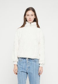 Biely flísový pulóver s vysokým golierom a zlatým zipsom. Mäkká textúra, rebrované manžety. Kombinovaný so svetlomodrými pásikavými rifľami s vysokým pásom.