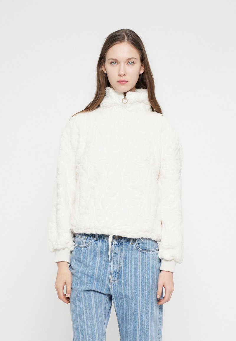 Biely flísový pulóver s vysokým golierom a zlatým zipsom. Mäkká textúra, rebrované manžety. Kombinovaný so svetlomodrými pásikavými rifľami s vysokým pásom.