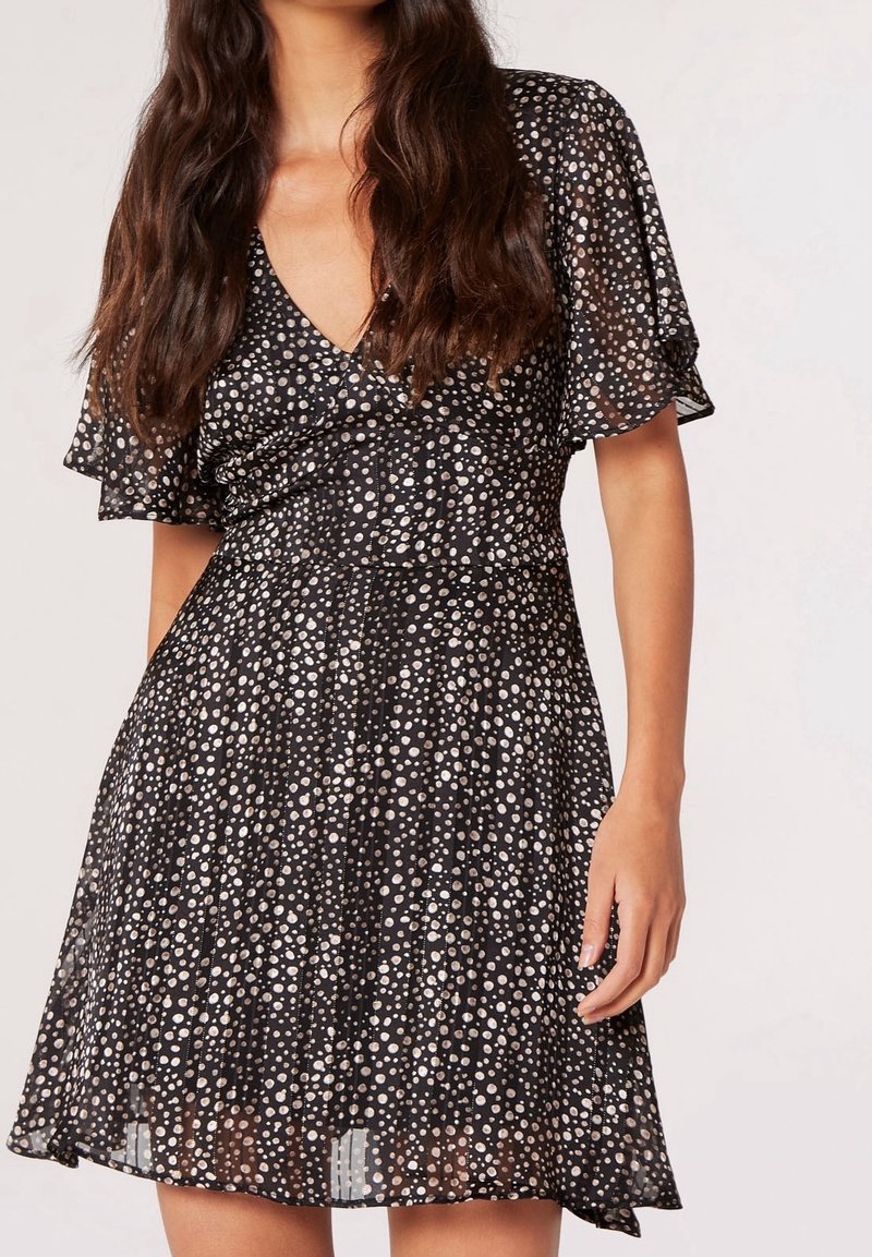 Femme portant une robe noire à pois blancs, manches volantées et un décolleté en V, debout devant un fond clair uni.