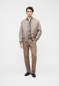 Veste bomber gris clair avec poignets côtelés, superposée sur un haut vert, associée à un pantalon marron à carreaux et des mocassins foncés. Design simple et moderne.
