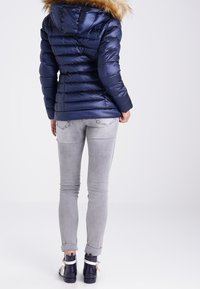 Doudoune bleue marine avec capuche en fausse fourrure, design matelassé, et jeans skinny bleu-gris, associée à des bottines sombres avec des accents blancs.