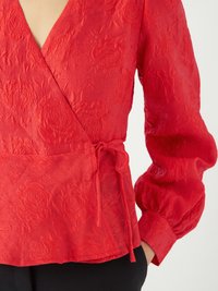 Blouse rouge texturée avec motif floral, décolleté en V profond, longues manches bouffantes et détail de nœud à la taille.