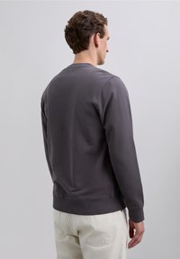 Bărbat cu păr castaniu, îmbrăcat într-o cămașă cu mânecă lungă de culoare gri închis și pantaloni bej deschis, stând cu fața spre spate, pe un fundal simplu.