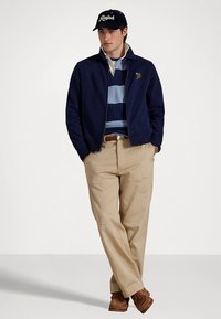 Casaco azul-marinho com fecho éclair por cima de uma camisa polo às riscas azuis, calças bege, sapatos castanhos e um boné azul-marinho com um logótipo branco. Roupa casual com um corte moderno.