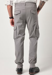 Szare spodnie cargo z elastycznymi mankietami i pasem, z wieloma kieszeniami oraz gładką fakturą materiału. Noszone z czarnymi butami.