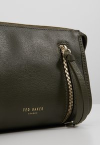 Borsa a mano in pelle verde oliva con cerniera dorata, caratterizzata da una superficie testurizzata e con un logo in rilievo dorato sulla parte anteriore.