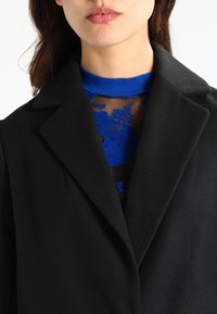 Sort frakke med reverskrave, der har en blå turtleneck under. Frakken ser skræddersyet ud med en glat tekstur.
