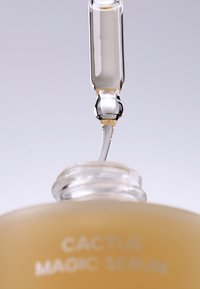 Sérum Magique au Cactus dans une bouteille en verre ambre ronde avec un compte-gouttes transparent, laissant échapper une goutte de sérum jaune clair.