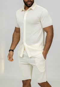 Chemise légère beige clair à manches courtes côtelées avec un col boutonné, associée à un short assorti à longueur genou. Les deux pièces présentent des poches latérales.