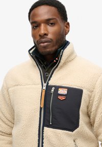 Beige Fleecejacke mit einem marineblauen Stoffpanel und einer Reißverschlusstasche auf der Brust. Verfügt über ein Logo-Patch und ein hohes Kragen-Design.