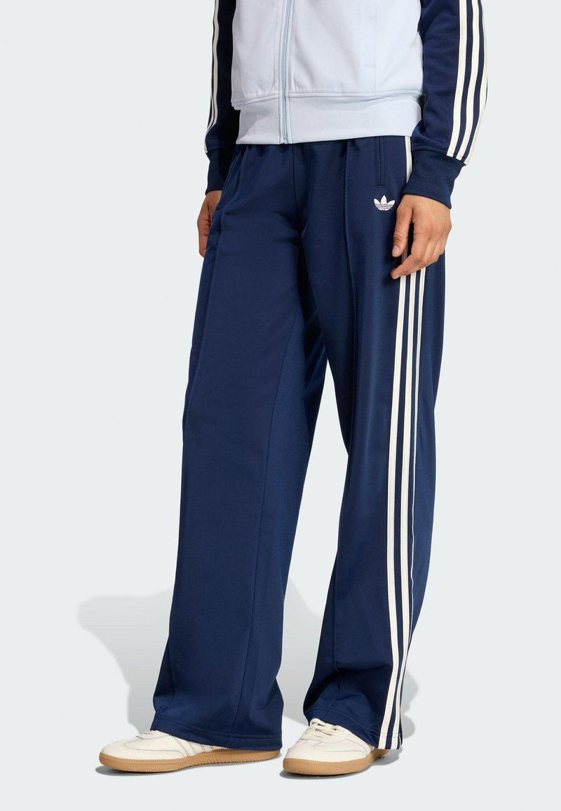 Pantalon de sport bleu marine avec un design à trois bandes blanches le long des côtés, confectionné en tissu lisse. Présente un logo brodé sur la cuisse gauche.