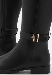Botas negras hasta la rodilla hechas de cuero sintético texturizado, con un acento de hebilla en tono dorado y una cremallera lateral para facilitar su uso.