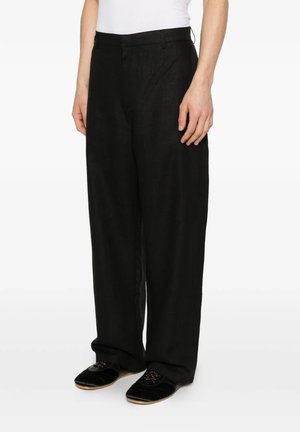Persona che indossa pantaloni neri larghi e scarpe slip-on in velluto nero con suola marrone chiaro, in piedi su uno sfondo bianco uniforme.