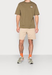 T-shirt à manches courtes vert olive avec le logo FILA, associée à un short beige et des baskets gris-vert avec des accents orange et des chaussettes.