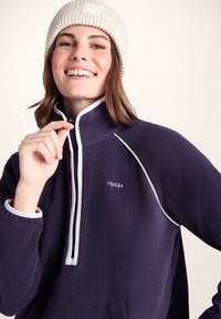 Smiling person wearing a white knit beanie and navy fleece jacket with white trim, pulling the front zipper up.
