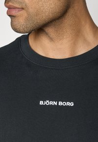 Črna bombažna majica s kratkimi rokavi in krožnim izrezom, na sprednji strani pa je bel vezen "BJÖRN BORG" logotip. Povečan pogled na izrez in teksturo blaga.