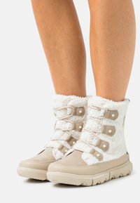 Beige ankelstøvler med hvit, luftig fôr, med snøring og brune detaljer. Teksturert stoff med en glatt gummisåle.