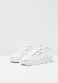 Baskets blanches à texture lisse, avec des accents dorés sur les œillets et le talon, et un logo discret sur le côté. Design plat à bout rond.