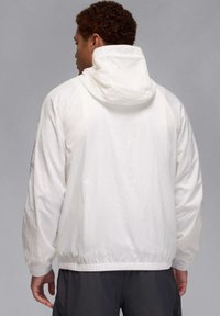 Chaqueta blanca con capucha, confeccionada en tela ligera, que presenta un dobladillo frontal, puños elásticos y un ajuste relajado con una textura suave.