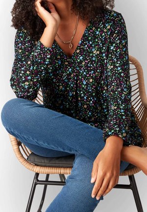 Frau mit lockigem Haar, die eine schwarze Bluse mit Blumenmuster und blaue Jeans trägt, sitzt mit überkreuzten Beinen auf einem Rattanstuhl mit schwarzem Kissen.