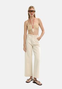 Kostium plażowy z bambusa z cienkimi ramiączkami, kremowe spodnie z szerokimi nogawkami z przednimi kieszeniami, czarne klapki, czerwona naszyjnik z wisiorkiem, oversize'owe okulary przeciwsłoneczne.